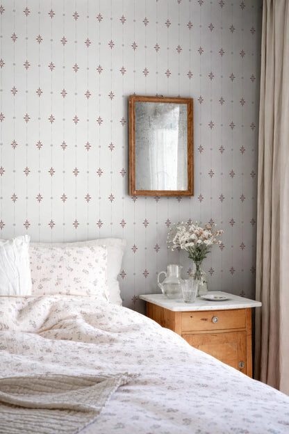 a cozy bedroom scene with a bed, a wooden nightstand, and a mirror on the wall. The bed is covered in a white comforter with a floral pattern, and there is a vase of flowers on the nightstand. The mirror is hung above the bed, and the wall behind it has a repeating pattern of small red and white stars.