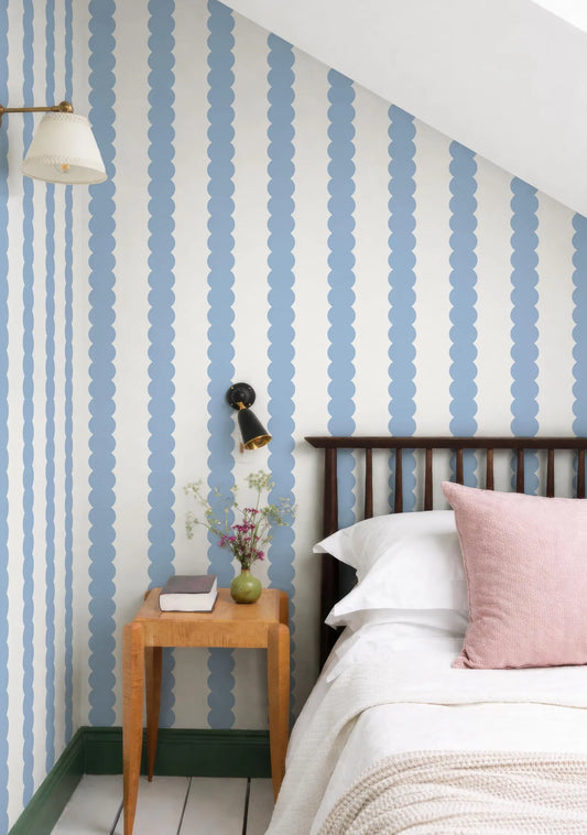 a cozy bedroom with a wooden bed frame, a wooden nightstand, and a lamp. The walls are covered in blue and white striped wallpaper, and the room has a slanted ceiling.