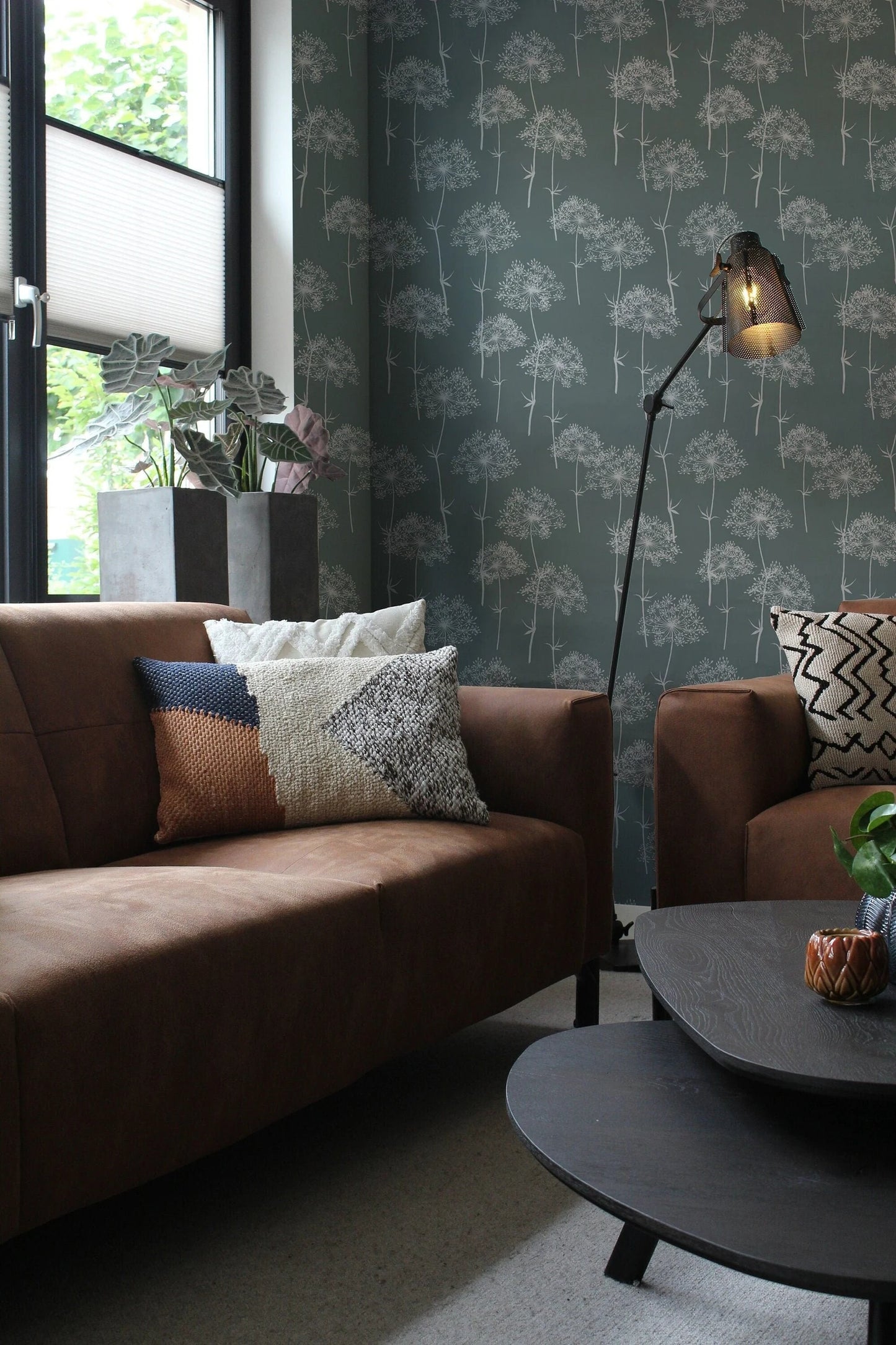 a cozy living room with a brown leather sofa, a wooden coffee table, and a floor lamp. The room has a patterned wallpaper and a window with blinds, allowing natural light to enter the space.