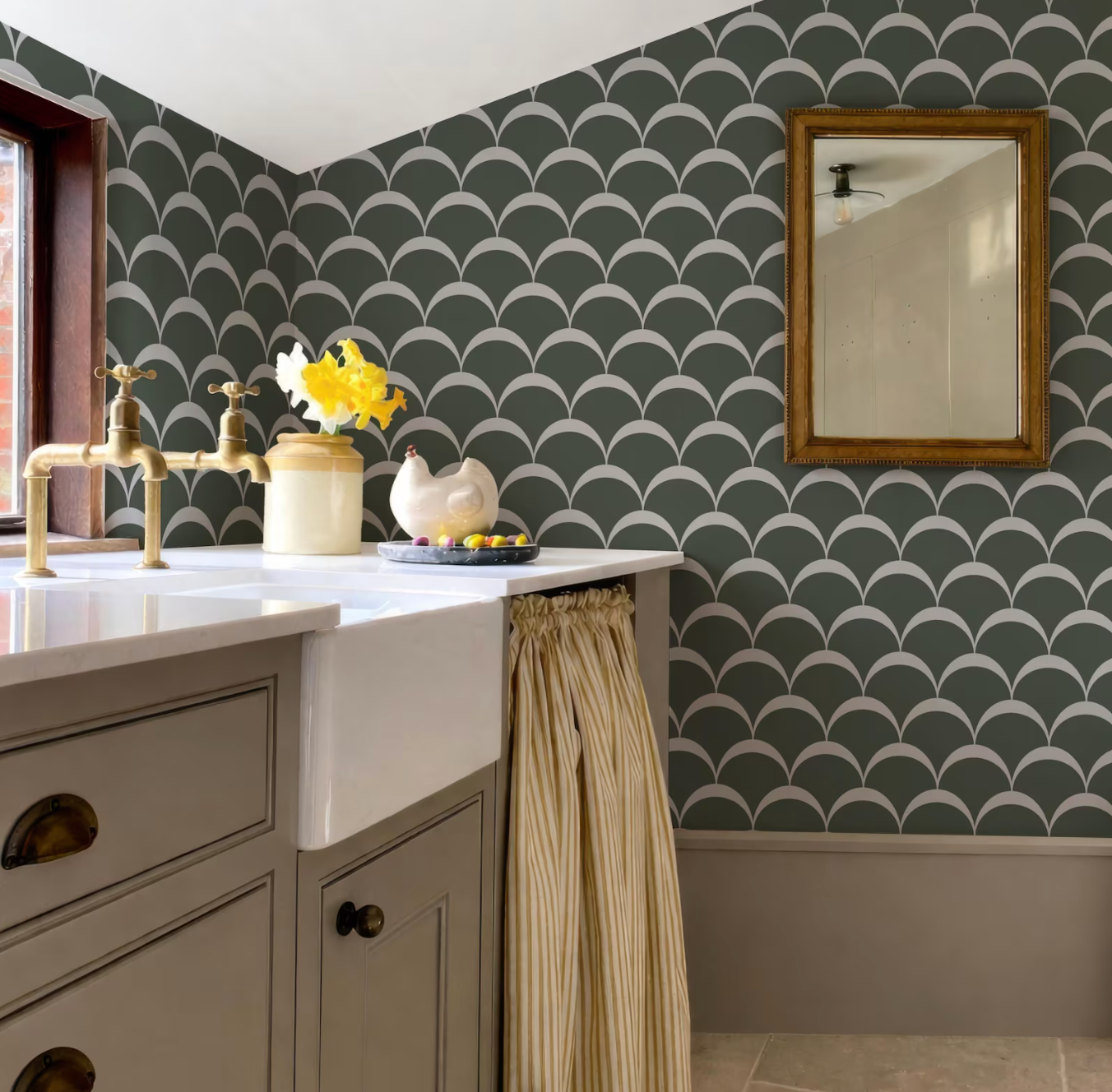 a bathroom with a white sink, a green and white patterned wallpaper, and a gold faucet. There is a small vase of yellow flowers on the counter, and a framed mirror is hanging on the wall.