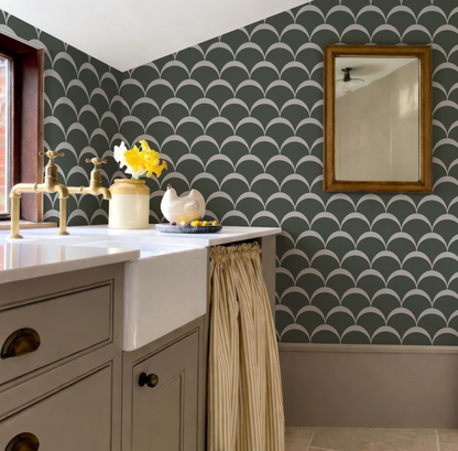 a bathroom with a white sink, a green and white patterned wallpaper, and a gold faucet. There is a small vase of yellow flowers on the counter, and a framed mirror is hanging on the wall.