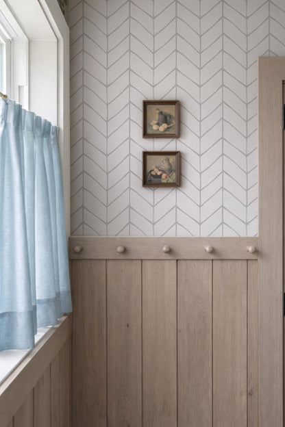 a room with a chevron-patterned wall, a wooden paneled wall, and a window with light blue curtains. There are two framed pictures hanging on the wall above the wooden paneling.