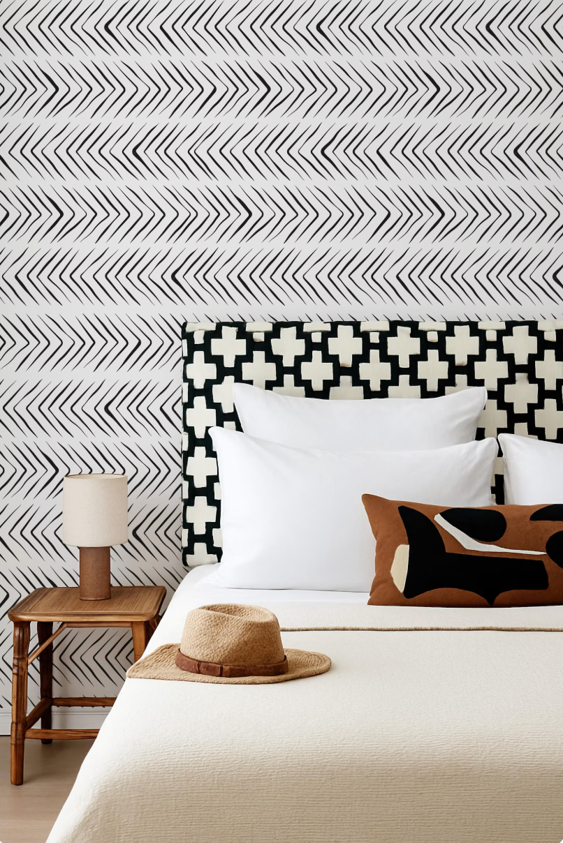 a bedroom with a white bed, a wooden nightstand, and a patterned headboard. A hat is placed on the bed, and there is a lamp on the nightstand.