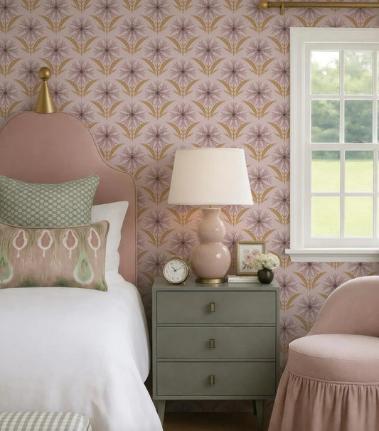 a bedroom with a pink floral wallpaper, a white bed with a green and white patterned pillow, a gray dresser with a lamp, and a pink armchair.