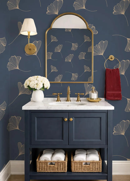 a bathroom vanity with a blue cabinet, a white countertop, and a gold faucet. Above the vanity is a gold-framed mirror with a gold leaf pattern. The wall behind the vanity has a blue and gold floral wallpaper. A red towel is hanging on a gold towel ring, and there is a white vase with white flowers on the countertop.