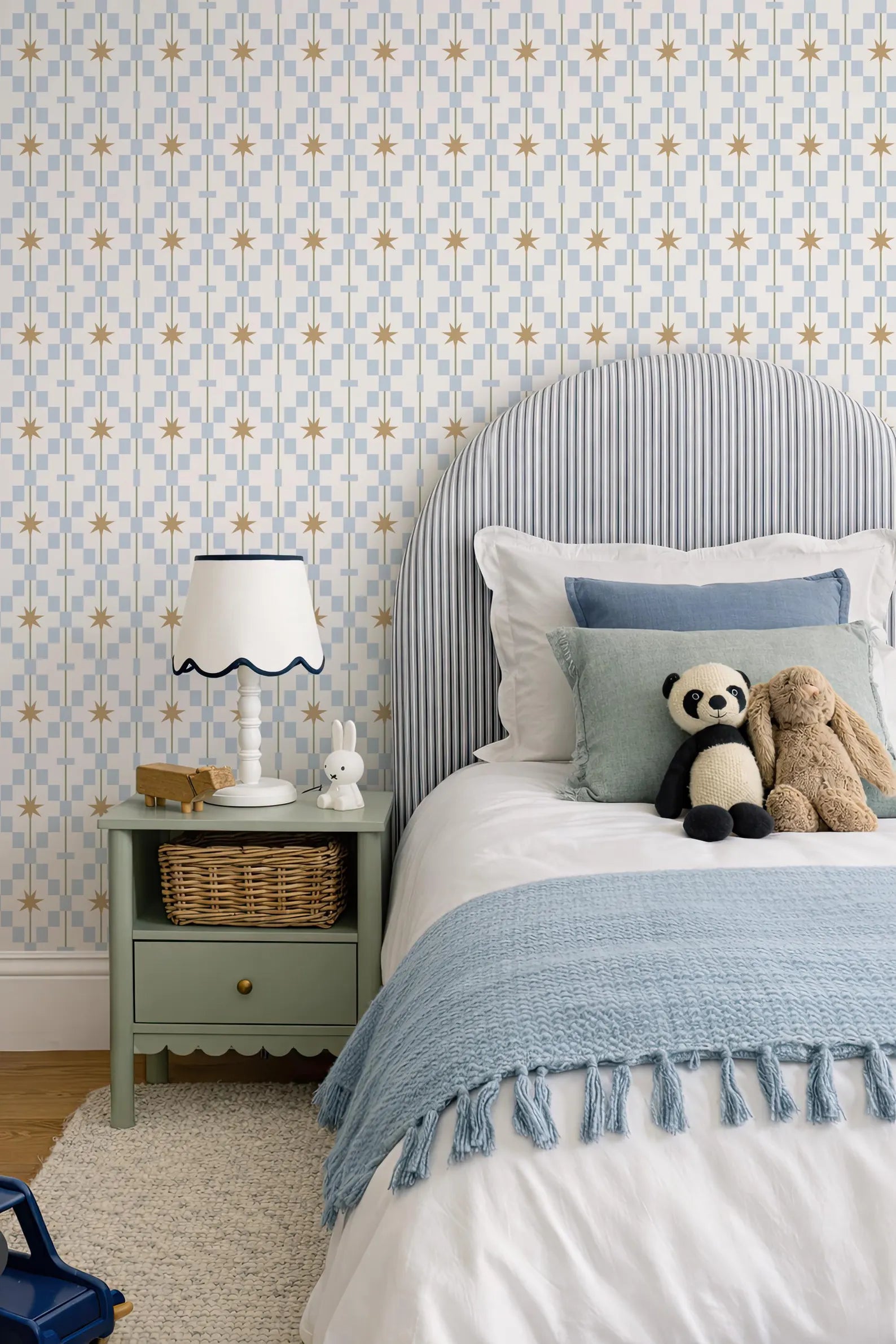 a cozy bedroom scene with a bed, nightstand, and stuffed animals. The bed has a blue and white striped headboard and a blue blanket. The nightstand has a white lamp and a small basket. There are two stuffed animals, a panda and a bunny, on the bed. The walls are covered in a patterned wallpaper with blue and gold stars.