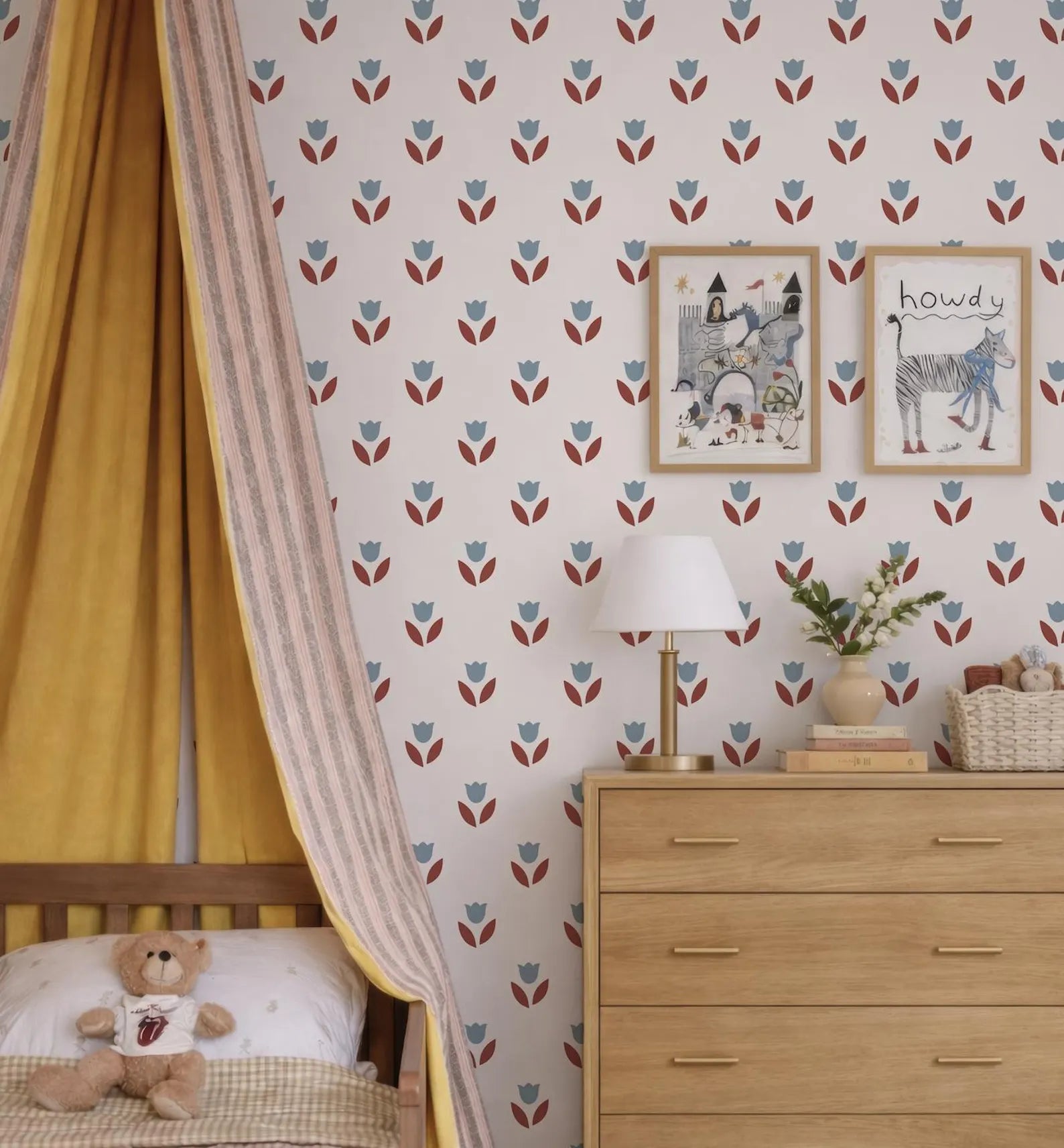 a cozy and charming bedroom scene, featuring a bed with a yellow and white striped bedspread, a wooden dresser with a lamp, and a wallpapered wall with a floral pattern. A teddy bear is also present in the room, adding a touch of warmth and playfulness to the space.