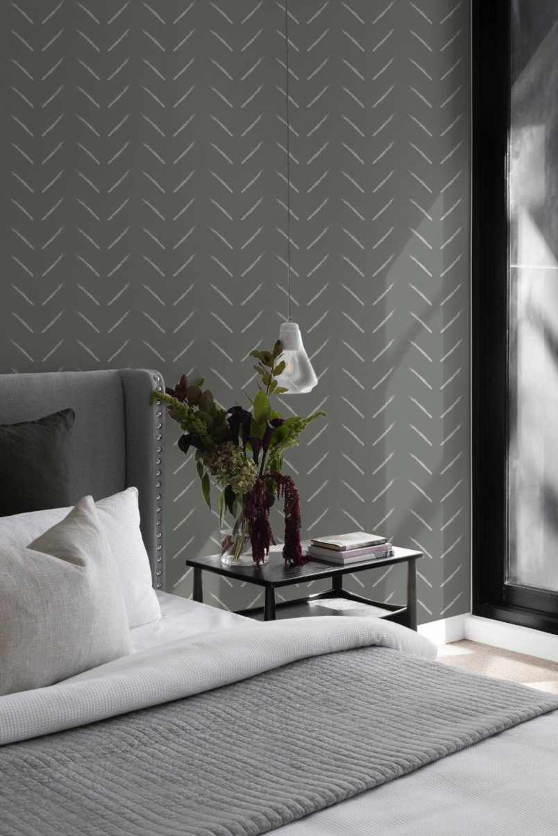 a modern bedroom with a gray and white color scheme. The focal point is a bed with a gray comforter and white pillows. On the bed, there is a nightstand with a vase of flowers and a lamp. The wall behind the bed has a gray and white chevron pattern, and the room is illuminated by natural light coming through a window.