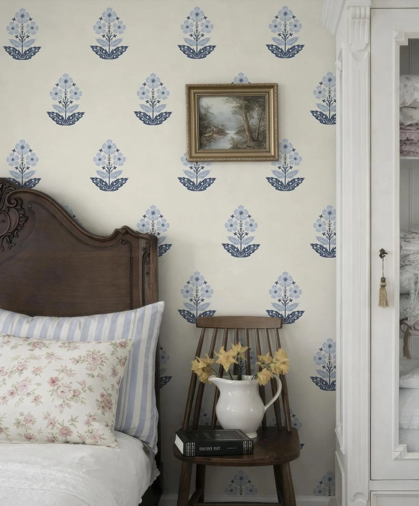 a bedroom with a wooden headboard, a white bedspread, and a wooden chair with a vase of yellow flowers. The wall behind the bed has a patterned wallpaper with blue flowers, and there is a framed picture hanging above the bed.