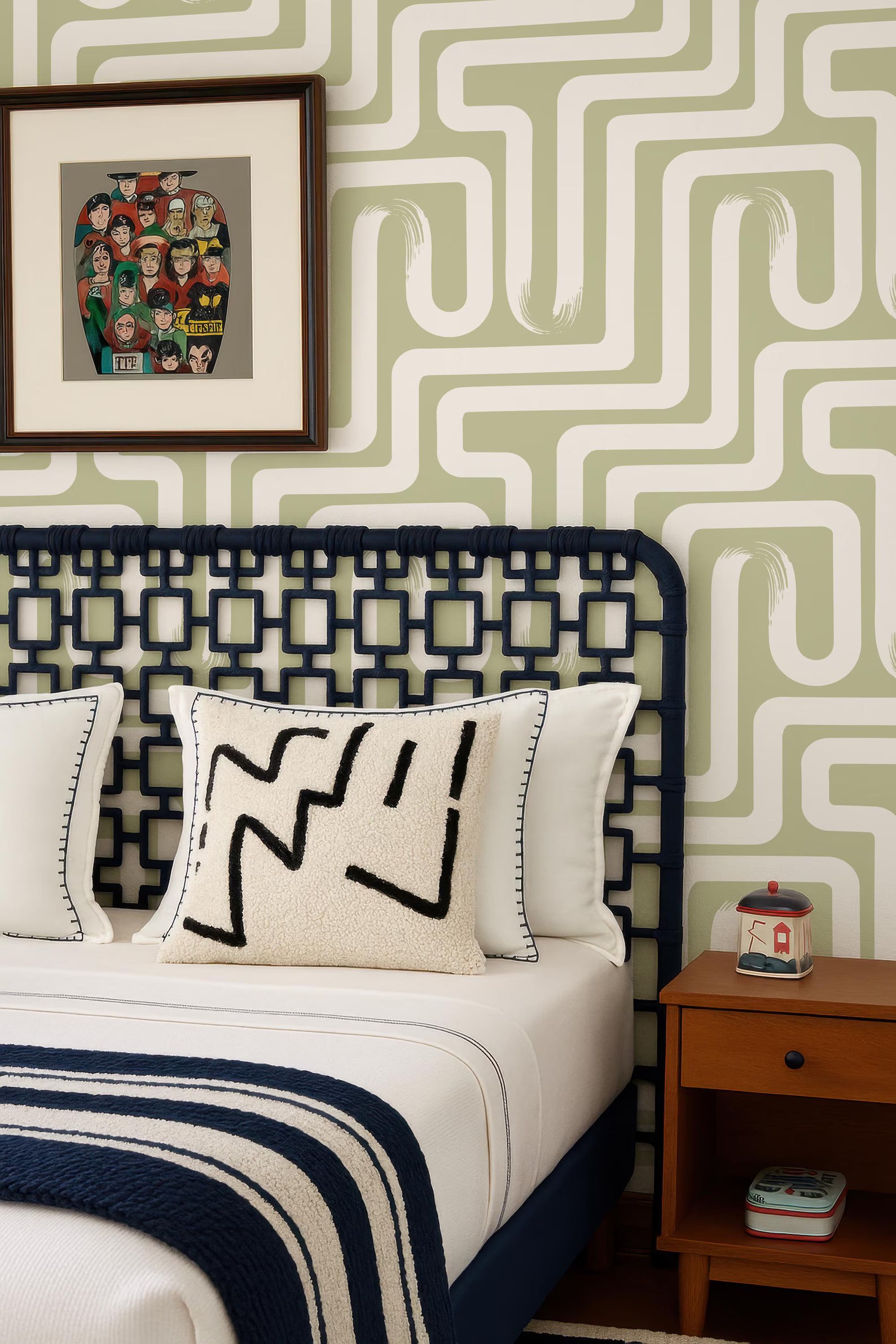 a bedroom with a bed, a framed artwork on the wall, and a wooden nightstand. The bed has a white comforter and a black and white patterned pillow. The wall has a green and white geometric pattern, and there is a small wooden table with a red and white container on it.