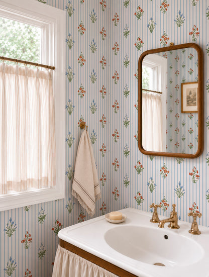 a bathroom with a white sink, a mirror, and a window with curtains. The walls are covered in a floral wallpaper pattern, and there is a towel hanging on the wall.