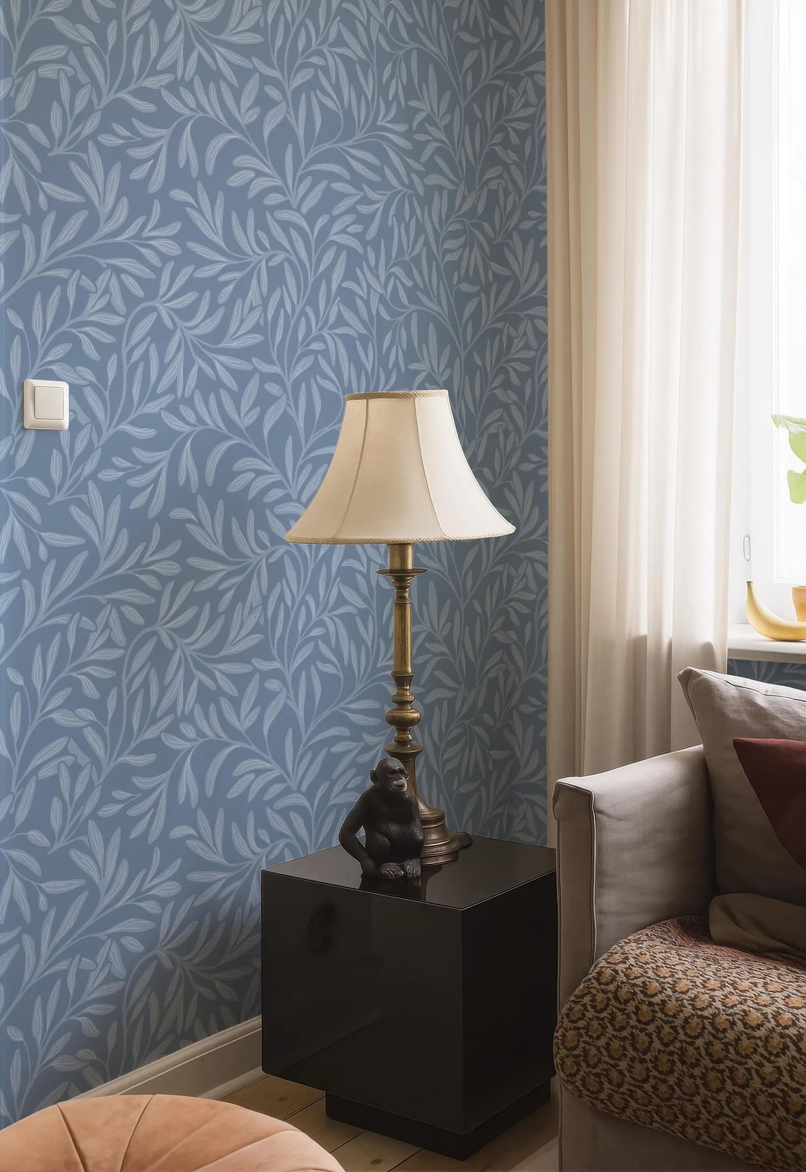 a living room with a blue leaf-patterned wallpaper, a white lamp with a gold base, a black side table, a beige sofa, and a brown and white patterned throw blanket.