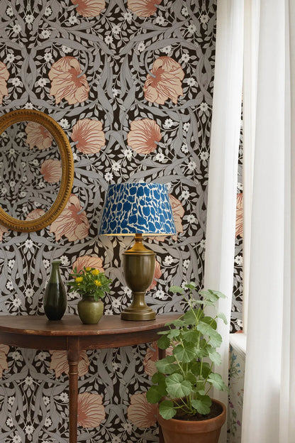 a table with a lamp, a vase with flowers, and a potted plant, set against a wall with a floral wallpaper pattern.