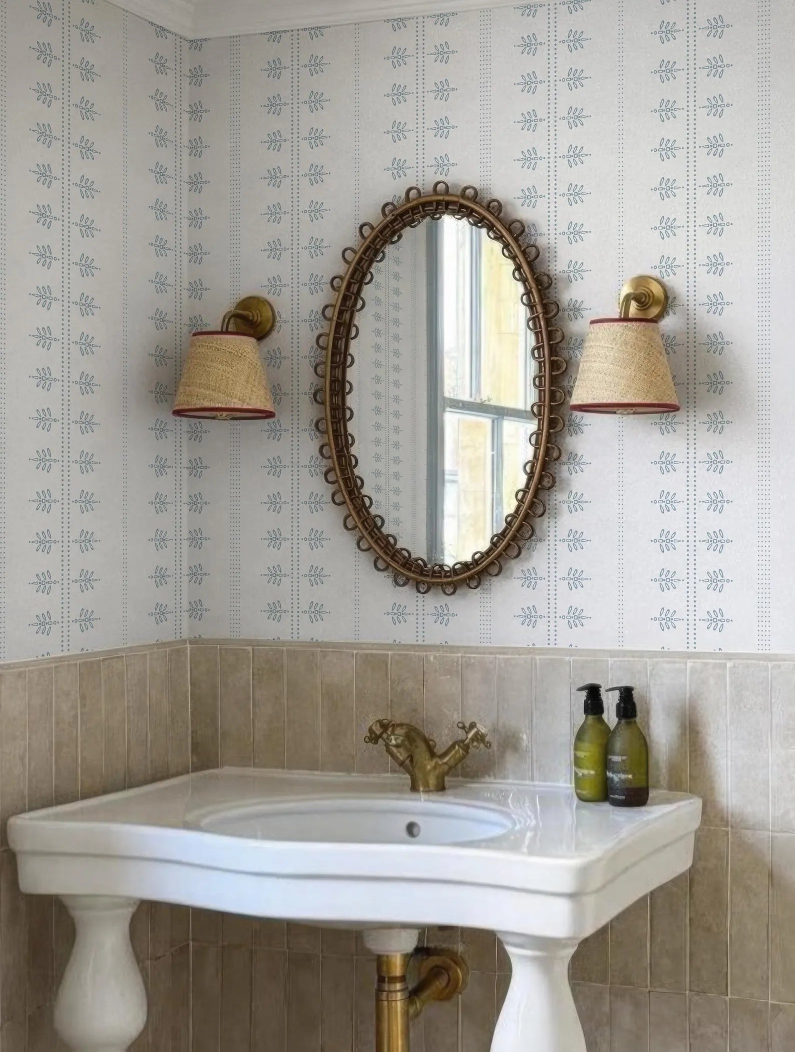 a bathroom with a white sink, a gold faucet, and a large oval mirror with a decorative frame. There are two wall sconces on either side of the mirror, and two bottles of soap are placed on the sink. The overall color scheme of the bathroom is white and gold, with a patterned wallpaper adding visual interest to the space.