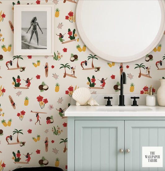 a bathroom with a light blue cabinet, a white sink, and a round mirror. The wall behind the cabinet is decorated with a tropical wallpaper pattern that includes various hula dancers, surfers, and other beach-themed elements. There is also a framed photo of a hula dancer on the wall.