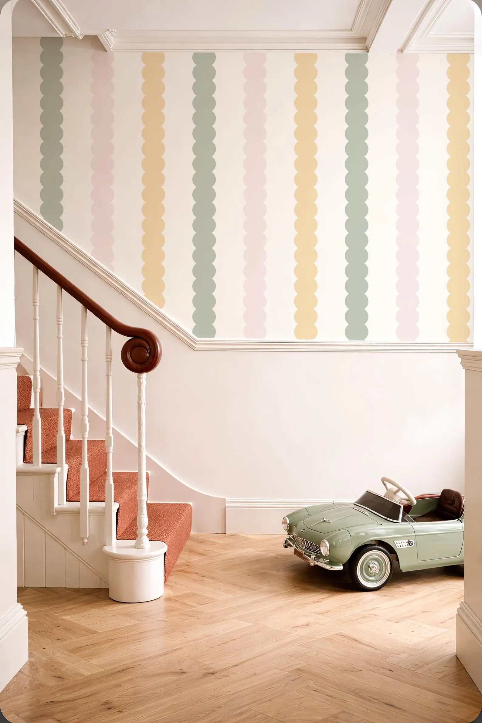 a vintage-style green toy car parked on a wooden floor next to a staircase with a white railing. The walls behind the car are decorated with colorful, vertical stripes in various pastel shades.
