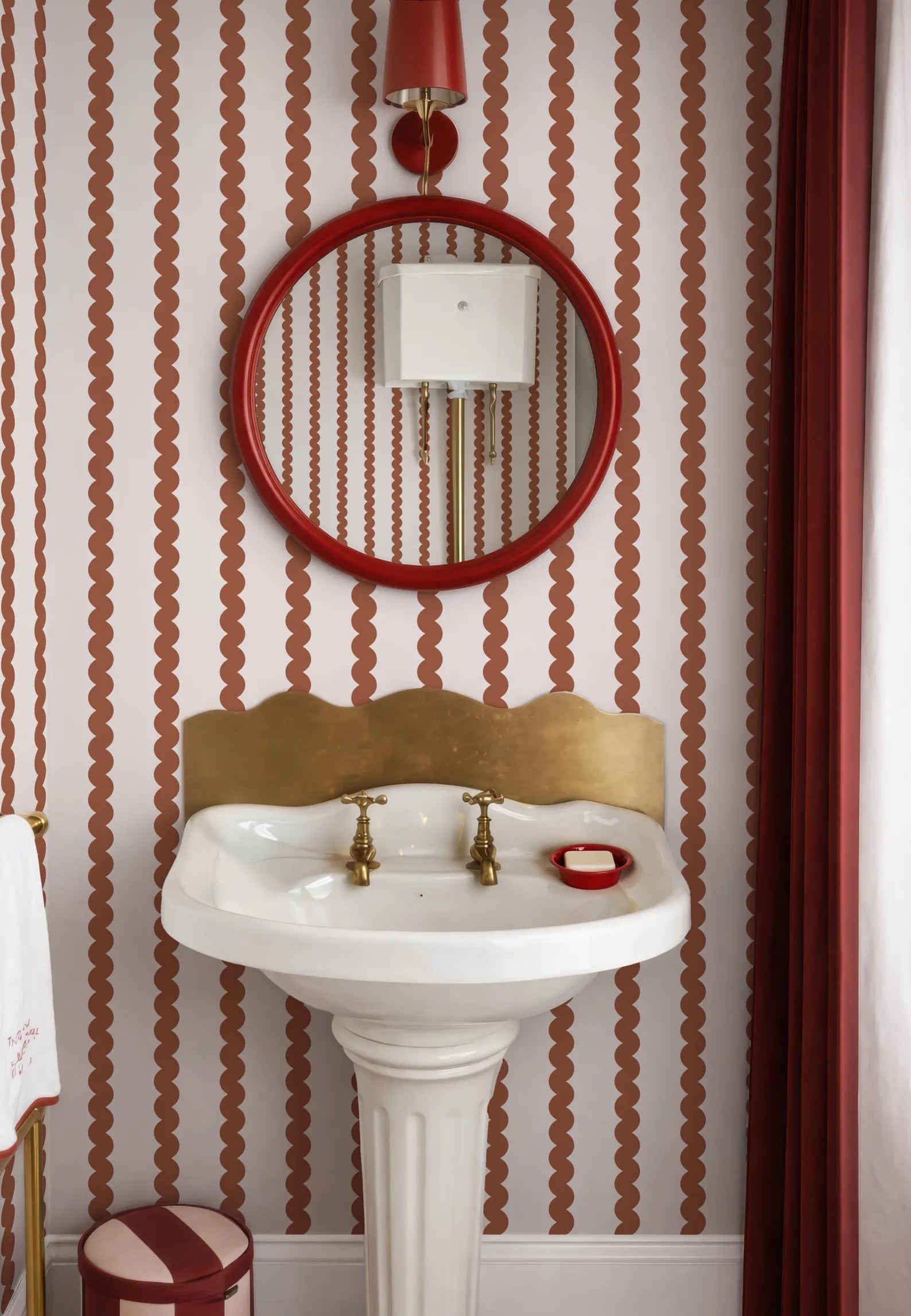 a bathroom with a white pedestal sink, a red circular mirror, and a gold faucet. The walls are covered in a red and white striped pattern, and there is a red curtain on the right side of the image.