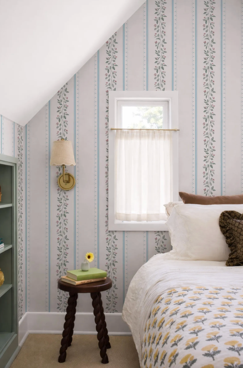 a cozy bedroom with a bed, a small wooden table, and a window with a white curtain. The walls are covered in a patterned wallpaper, and there is a lamp on the wall.