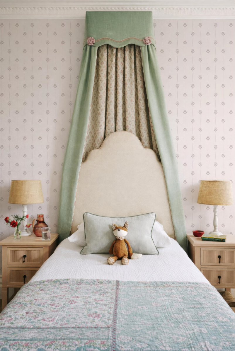 a cozy bedroom with a bed, nightstands, and a decorative canopy. The bed has a white comforter and a stuffed animal on it. The nightstands have lamps on them, and the canopy is green and has a patterned design. The walls are covered in wallpaper with a repeating pattern.