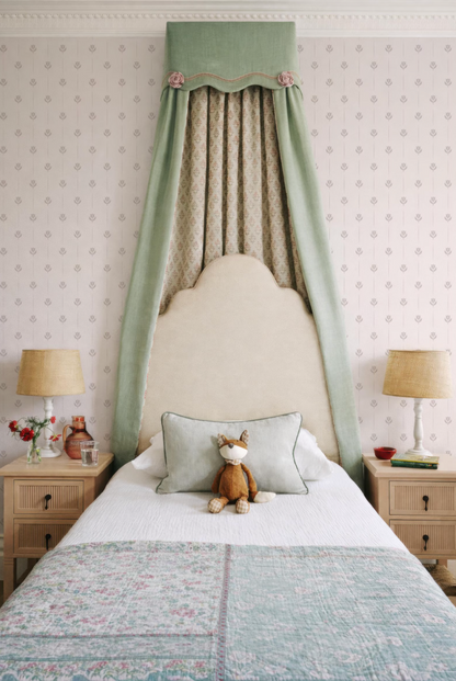 a cozy bedroom with a bed, nightstands, and a decorative canopy. The bed has a white comforter and a stuffed animal on it. The nightstands have lamps on them, and the canopy is green and has a patterned design. The walls are covered in wallpaper with a repeating pattern.