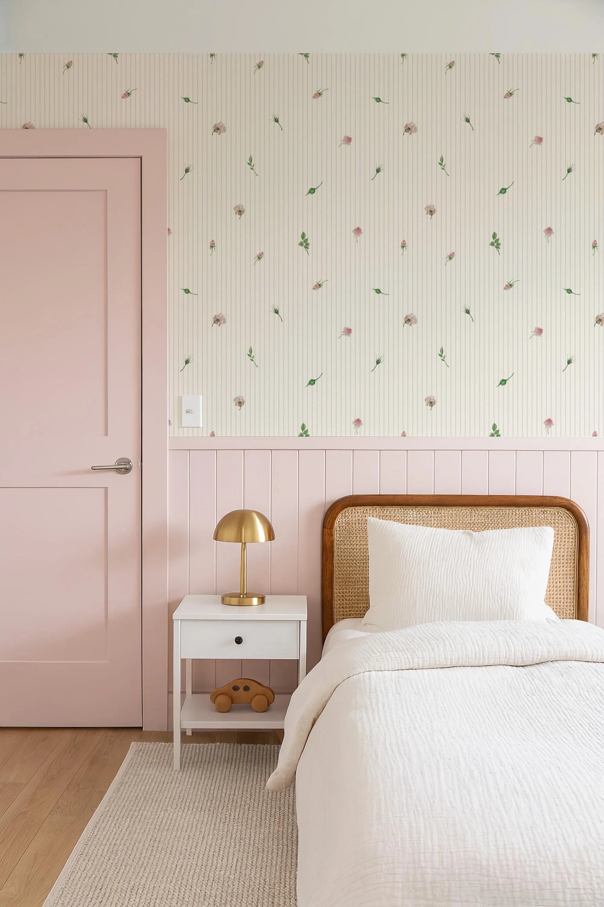 a cozy bedroom with a pink door, a white bed with a wooden headboard, and a white nightstand with a lamp. The walls are covered in a floral wallpaper, and the overall color scheme is predominantly pink and white.