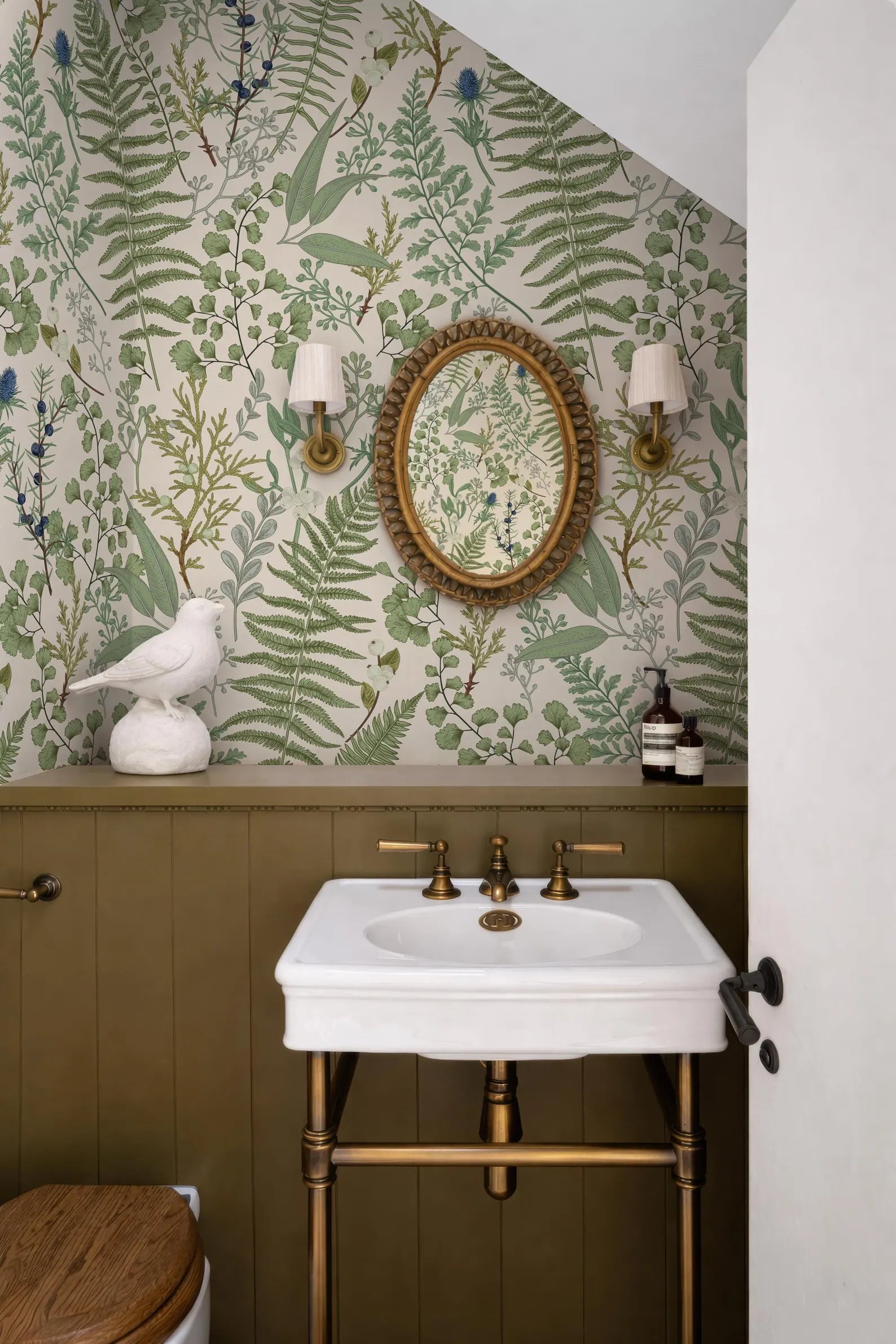 a bathroom with a white sink, a round mirror, and a bird figurine on a shelf. The walls are covered in a green and white floral wallpaper, and there are two wall sconces above the mirror.