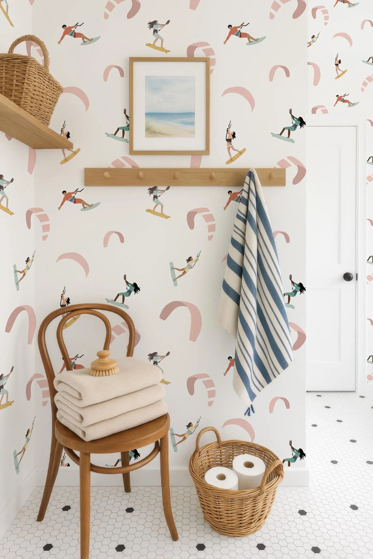 a bathroom with a wooden chair, a towel rack with a striped towel, and a basket of toilet paper. The walls are decorated with a pattern of surfers and kites, creating a beach-themed atmosphere.