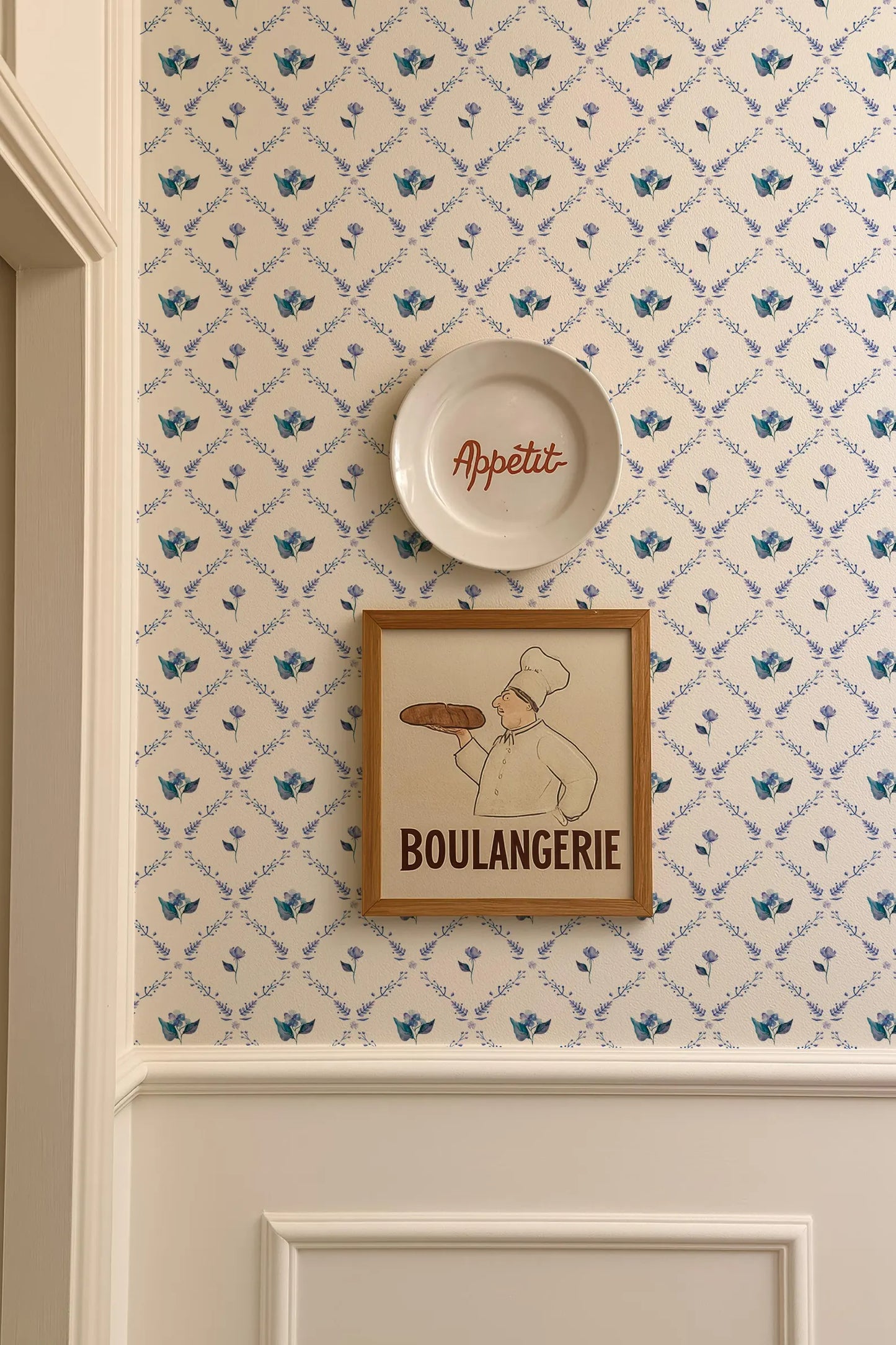 a wall with a framed picture of a chef holding a loaf of bread, and a plate with the word "Appétit" written on it. The wall has a patterned wallpaper with blue flowers.