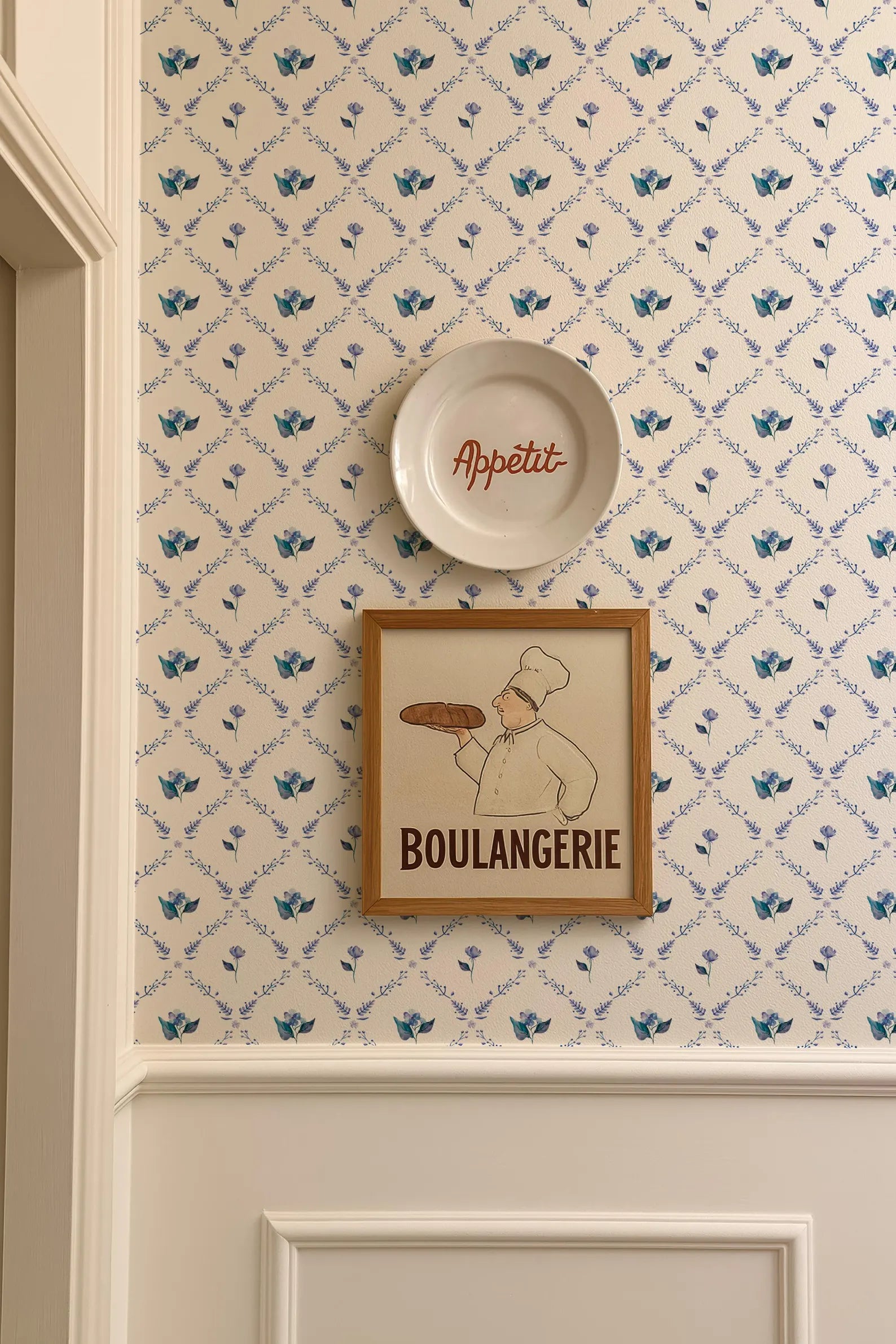 a wall with a framed picture of a chef holding a loaf of bread, and a plate with the word "Appétit" written on it. The wall has a patterned wallpaper with blue flowers.