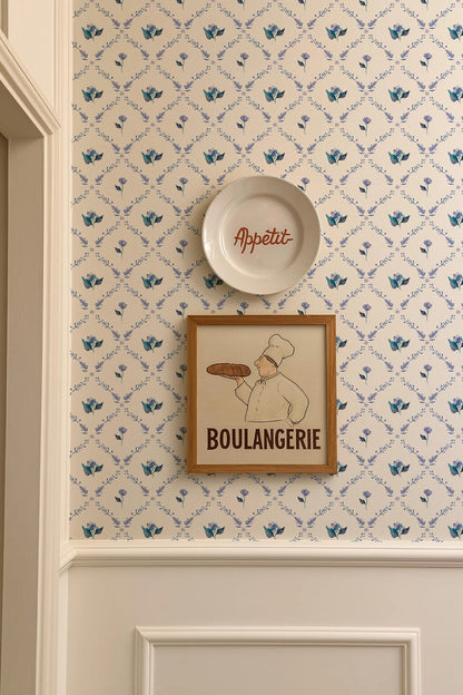 a wall with a framed picture of a chef holding a loaf of bread, and a plate with the word "Appétit" written on it. The wall has a patterned wallpaper with blue flowers.