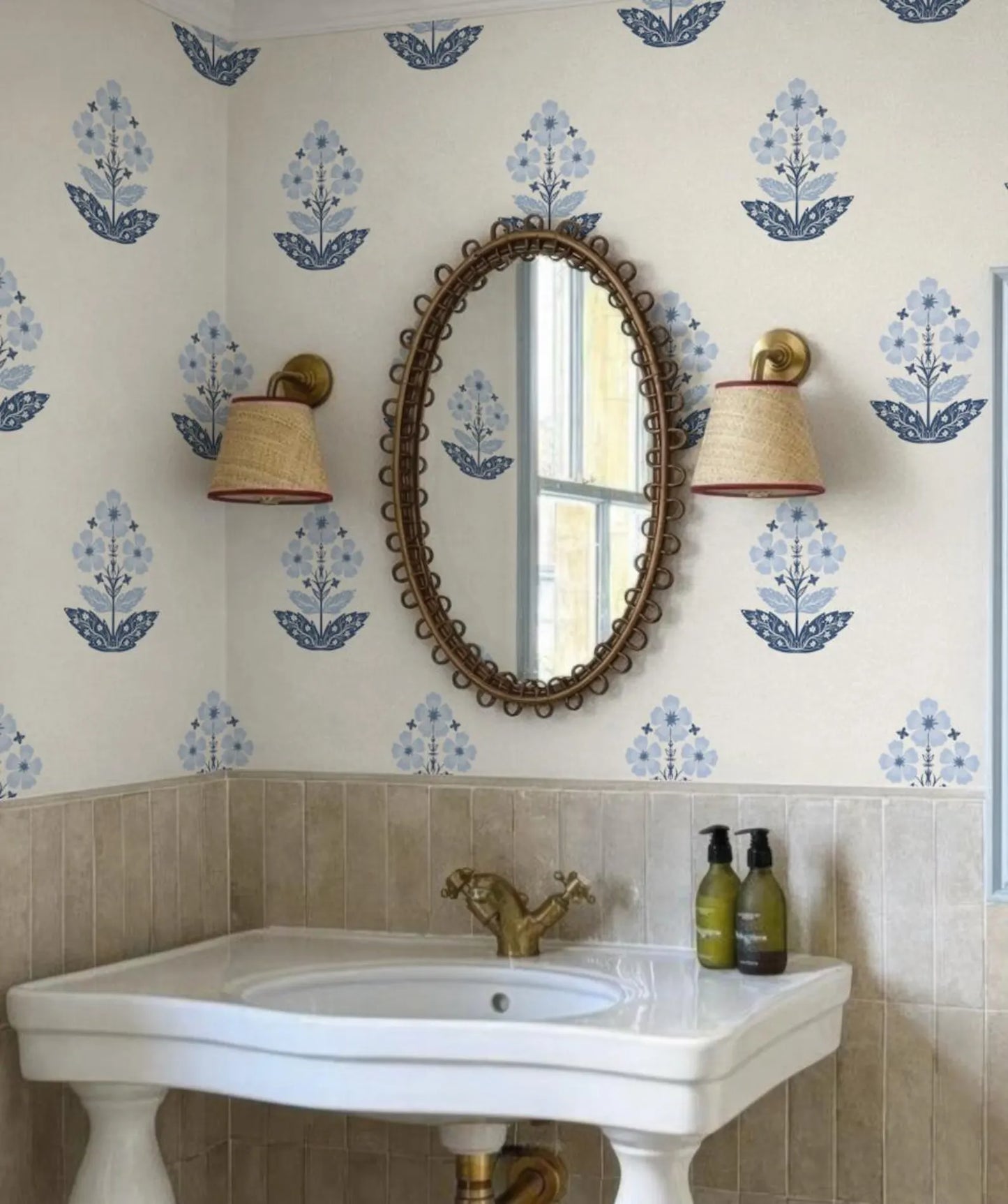 a bathroom with a white sink, a mirror, and two wall sconces. The walls are decorated with blue floral wallpaper, and there are two green bottles on the sink.