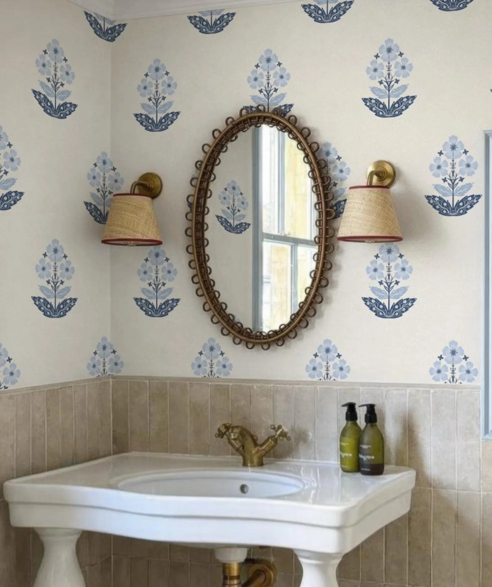 a bathroom with a white sink, a mirror, and two wall sconces. The walls are decorated with blue floral wallpaper, and there are two green bottles on the sink.