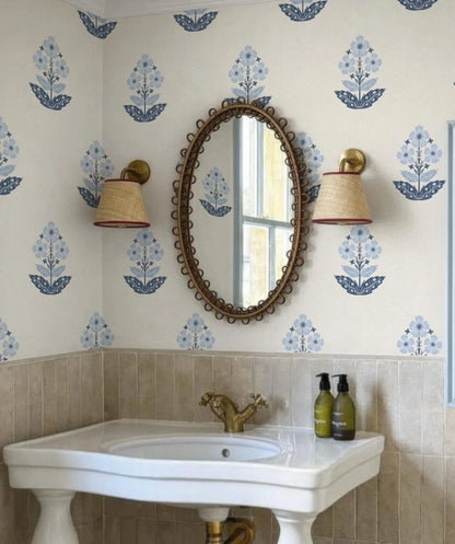 a bathroom with a white sink, a mirror, and two wall sconces. The walls are decorated with blue floral wallpaper, and there are two green bottles on the sink.
