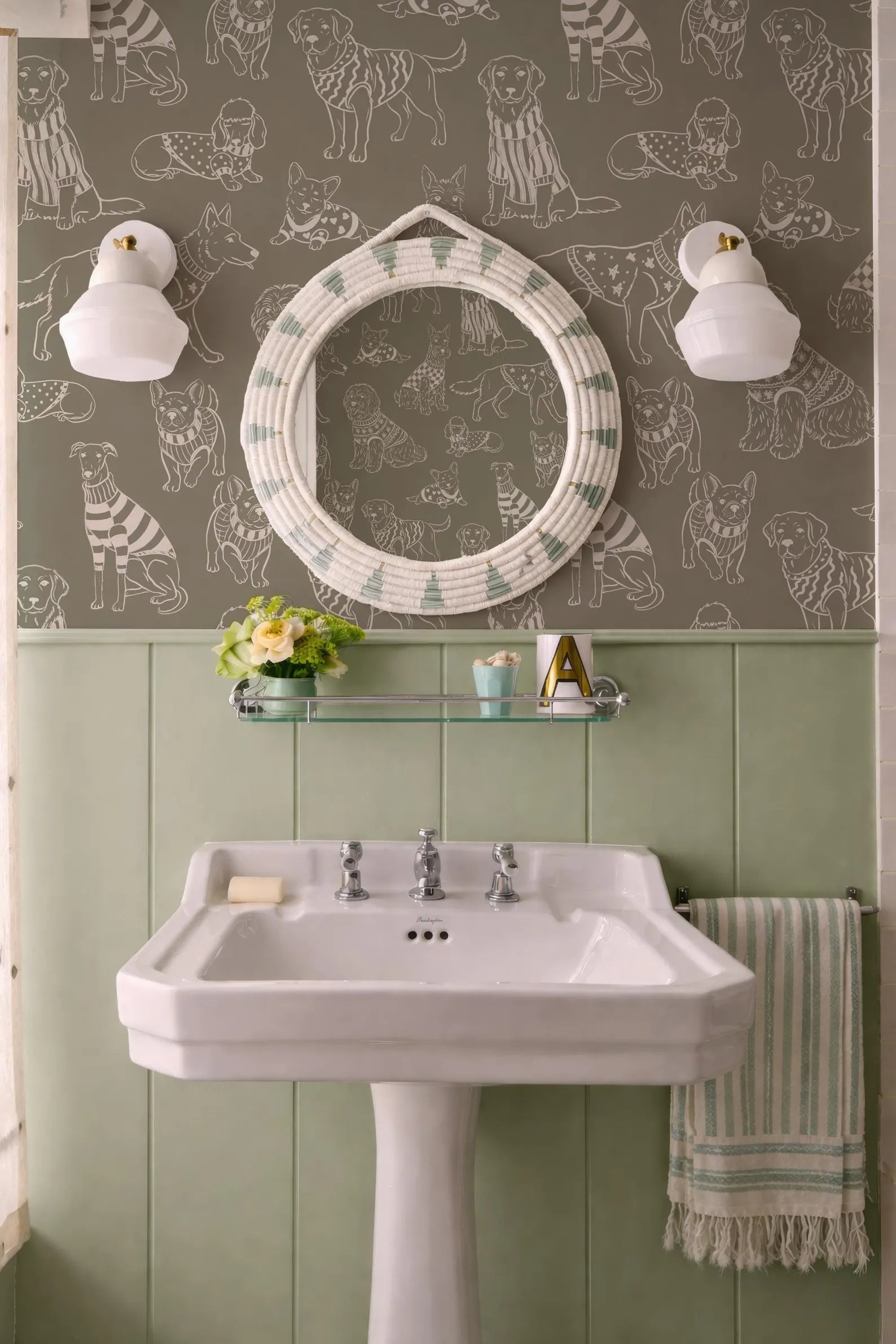 a bathroom with a white pedestal sink, a round mirror with a dog print frame, and a striped towel. The walls are covered in a patterned wallpaper with various dog illustrations.
