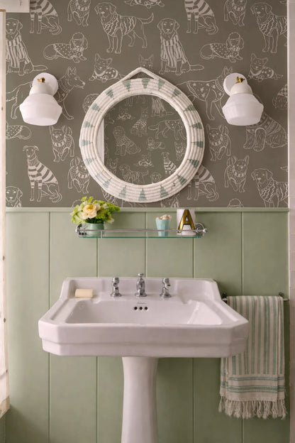 a bathroom with a white pedestal sink, a round mirror with a dog print frame, and a striped towel. The walls are covered in a patterned wallpaper with various dog illustrations.