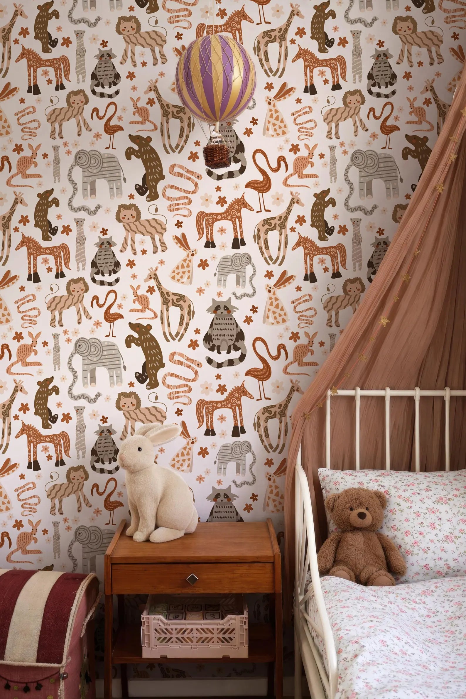 a child's bedroom with a white metal bed frame, a wooden nightstand, and a stuffed rabbit. The walls are covered in a patterned wallpaper featuring various animals and objects, including a hot air balloon.