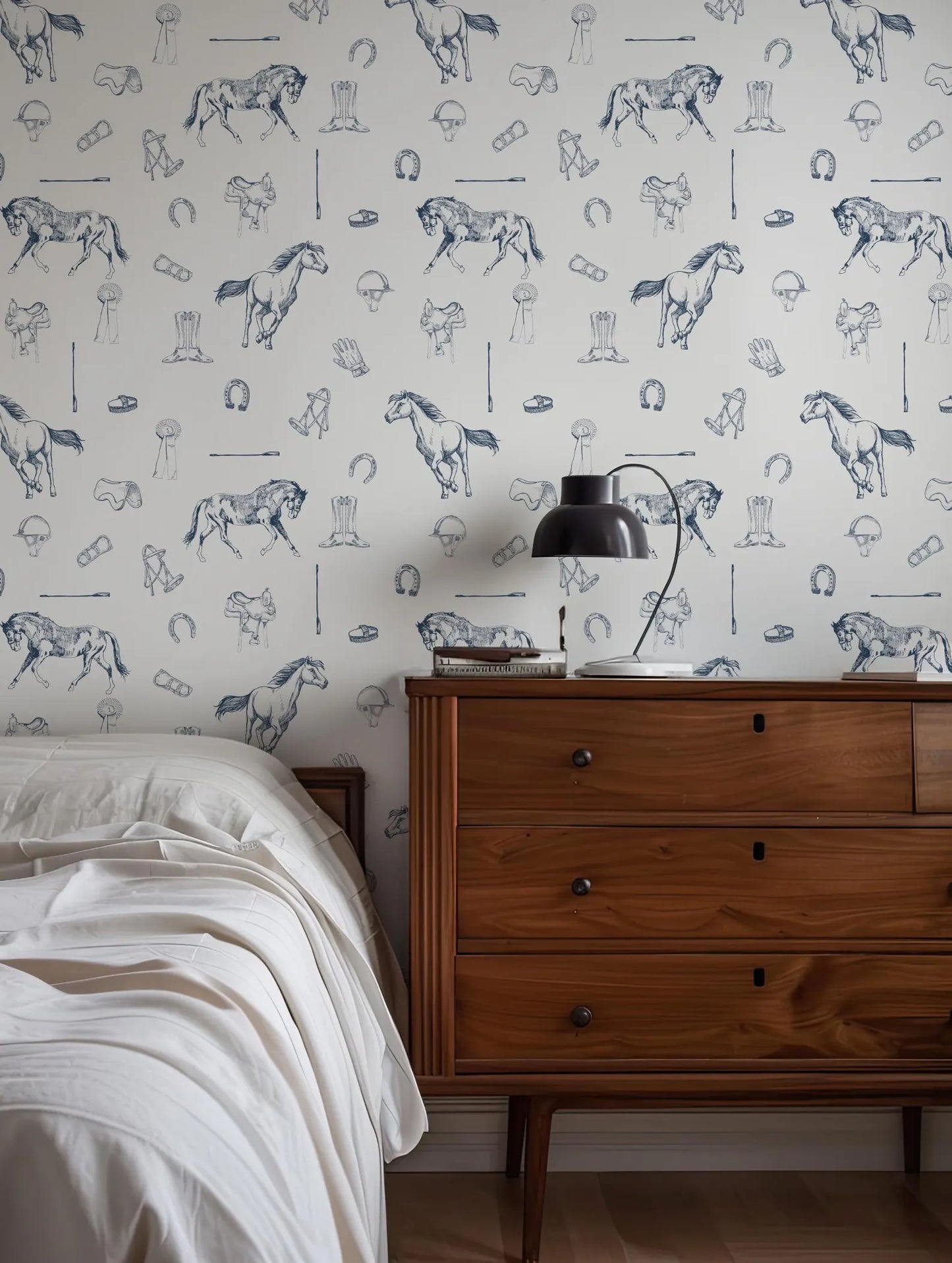 a bedroom setting with a wooden dresser and a bed. The dresser has a lamp on top, and the wall behind it has a wallpaper pattern of horses and other horse-related illustrations.