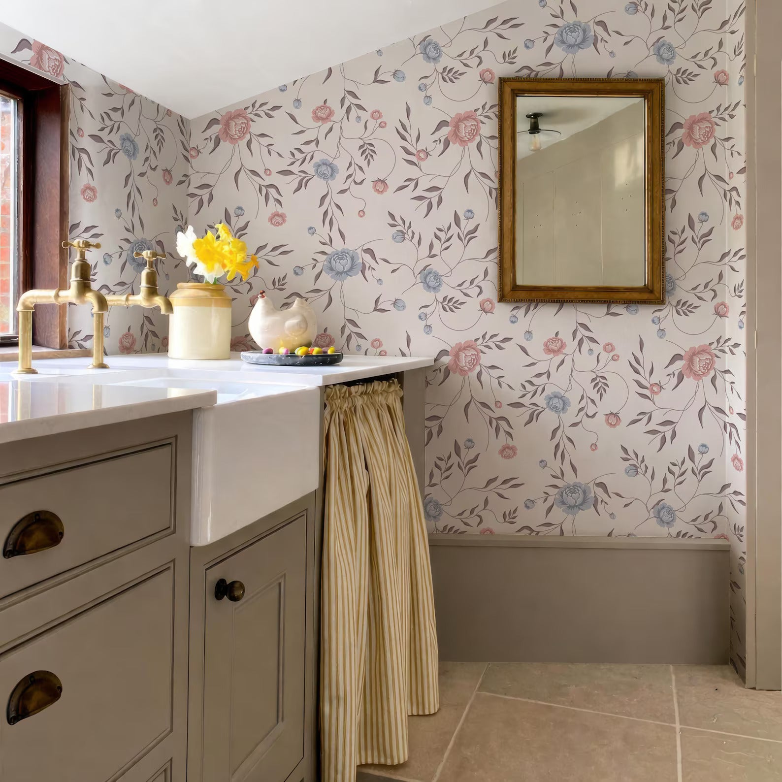 a bathroom with a floral wallpaper, a sink, and a mirror. The sink has a white basin and a vase of yellow flowers on it. The mirror is hanging on the wall above the sink. The overall color scheme of the bathroom is neutral, with a touch of yellow from the flowers.