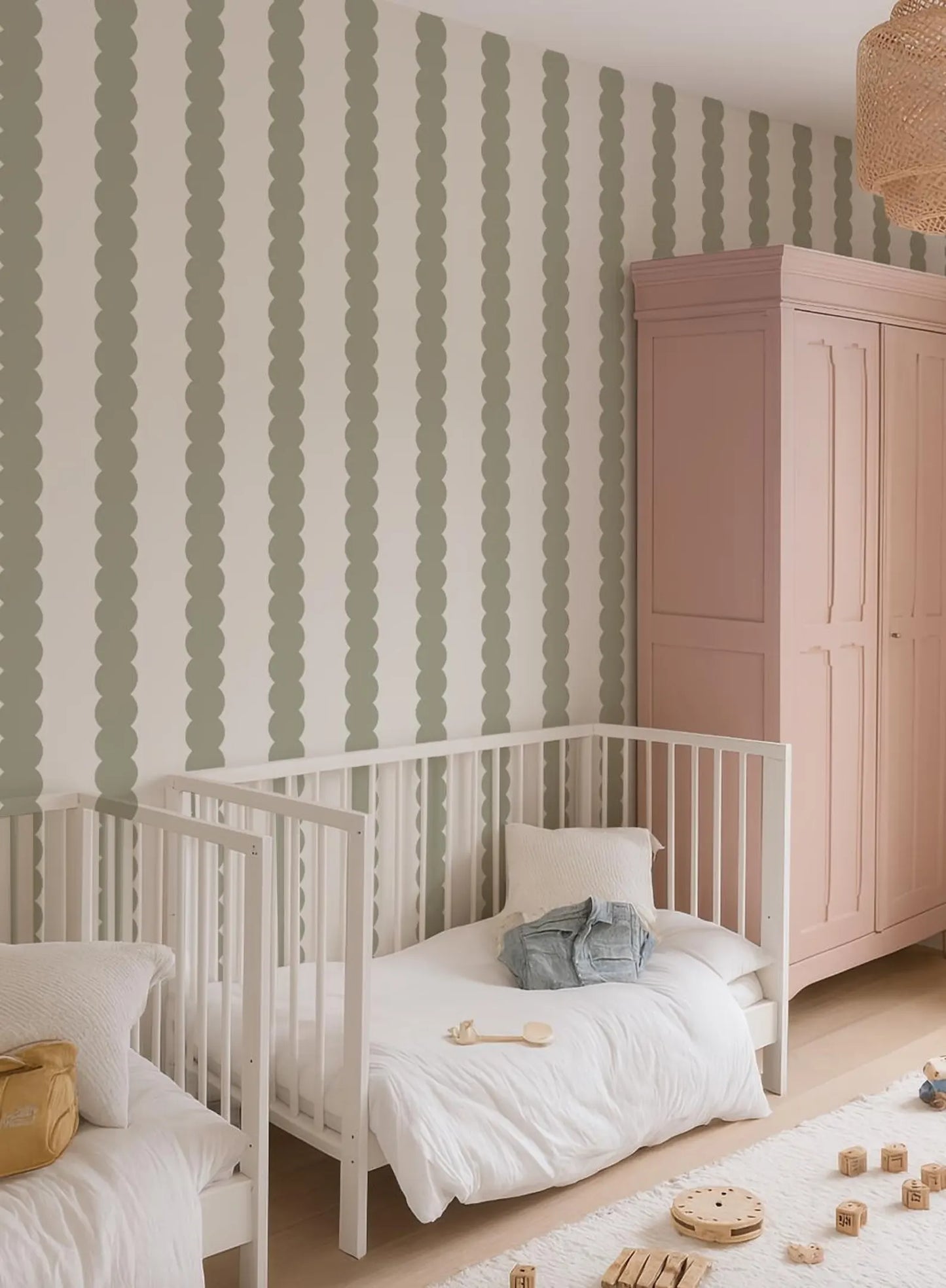 a cozy nursery with two white cribs, a pink armoire, and a green and white striped wallpaper. The cribs are positioned next to each other, and there are wooden blocks scattered on the floor.