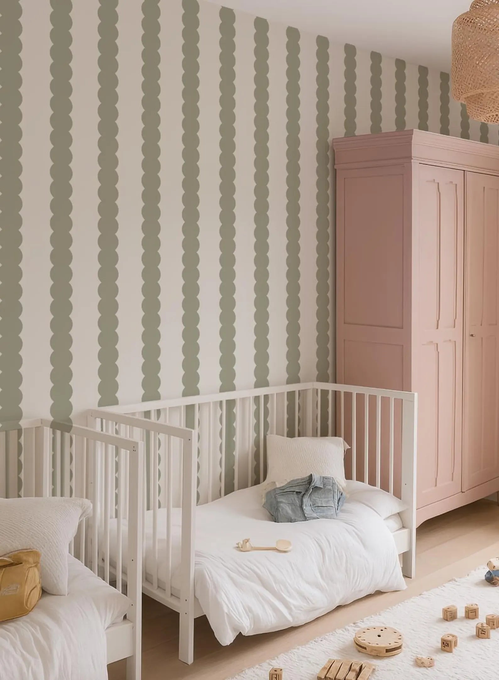 a cozy nursery with two white cribs, a pink armoire, and a green and white striped wallpaper. The cribs are positioned next to each other, and there are wooden blocks scattered on the floor.