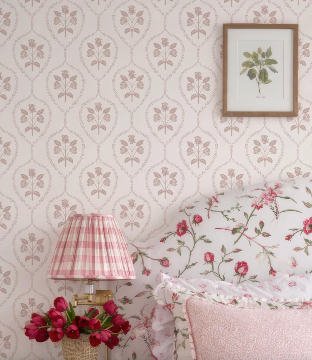 a bedroom setting with a floral print wallpaper, a lamp with a pink and white checkered lampshade, a framed picture of a plant, and a bed with a white and pink floral comforter and matching pillows.