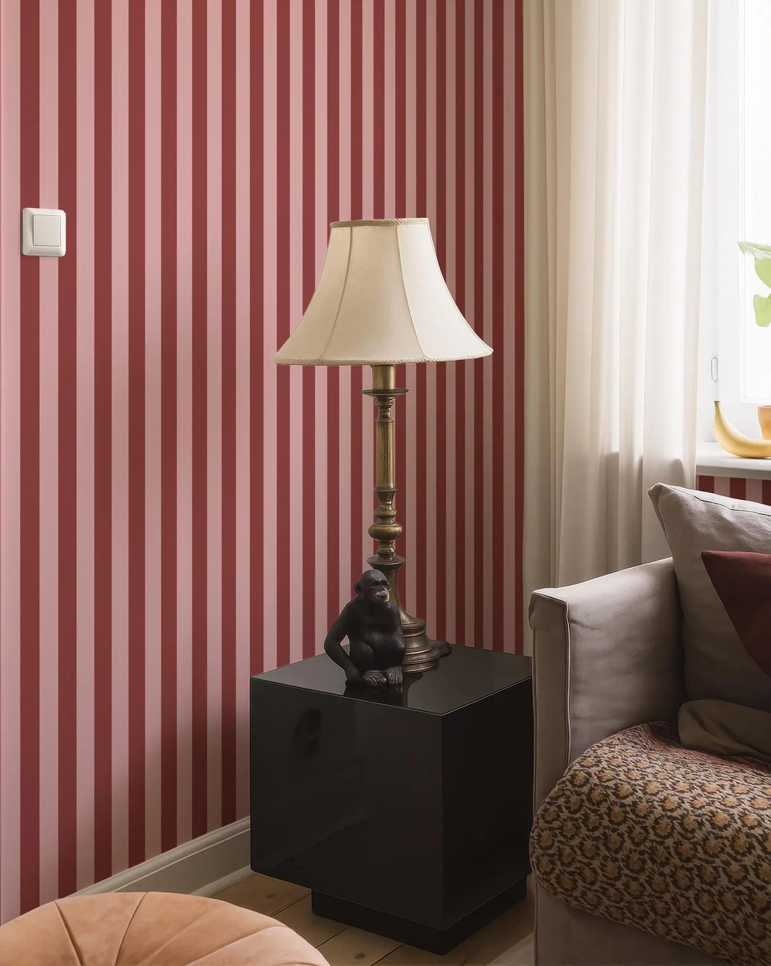 a living room with a red and white striped wallpaper, a black lamp on a black side table, a gray sofa, and a gold-colored vase on a windowsill.