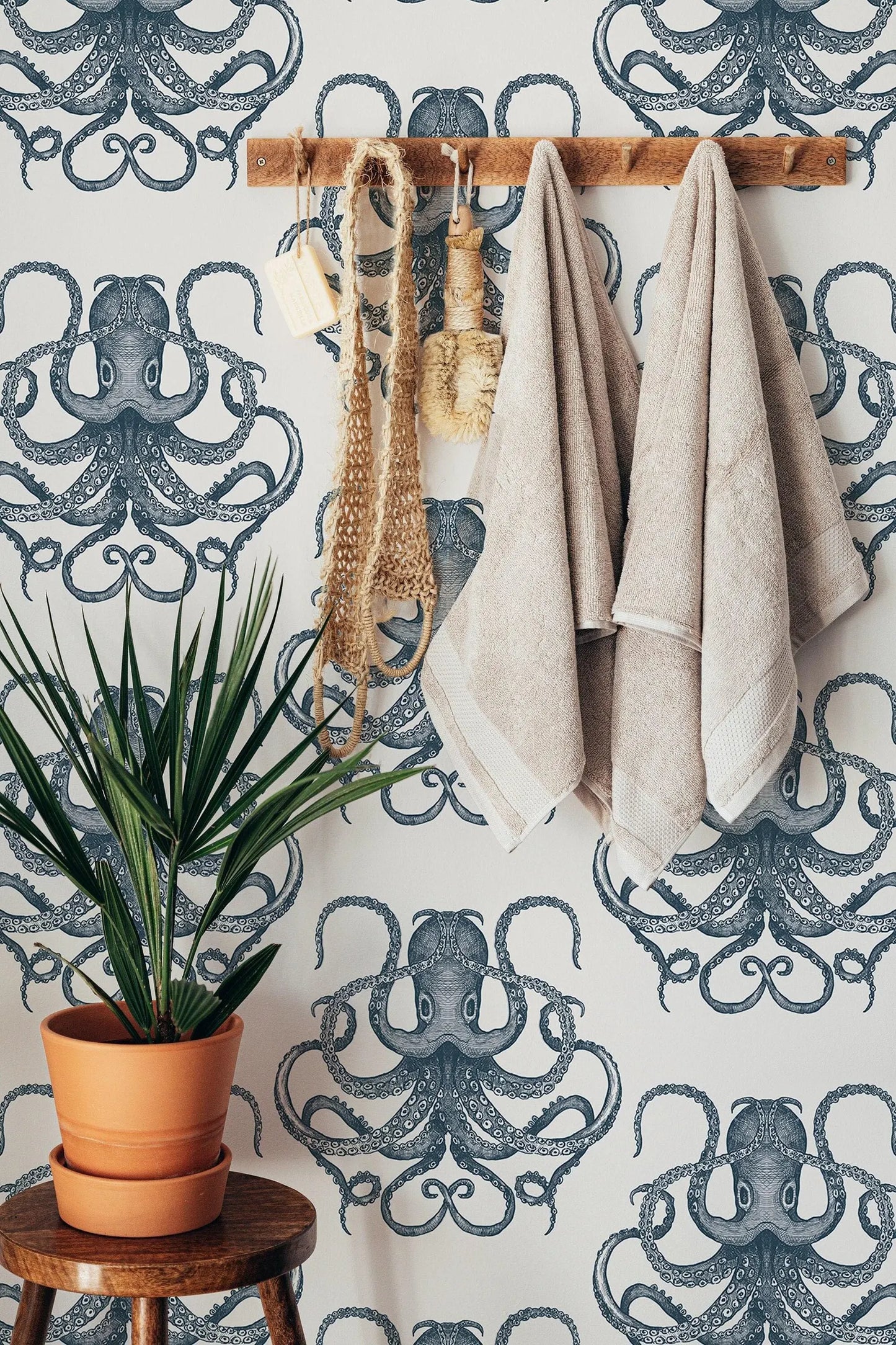 a bathroom setting with a wallpapered wall featuring a repeating pattern of octopus designs. On the wall, there are two towels hanging, a potted plant, and a wooden stool.
