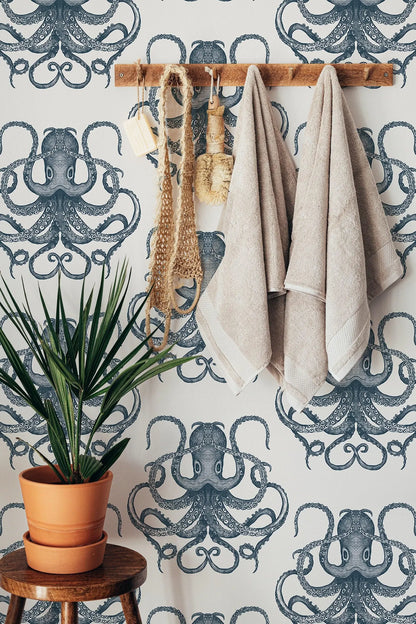 a bathroom setting with a wallpapered wall featuring a repeating pattern of octopus designs. On the wall, there are two towels hanging, a potted plant, and a wooden stool.