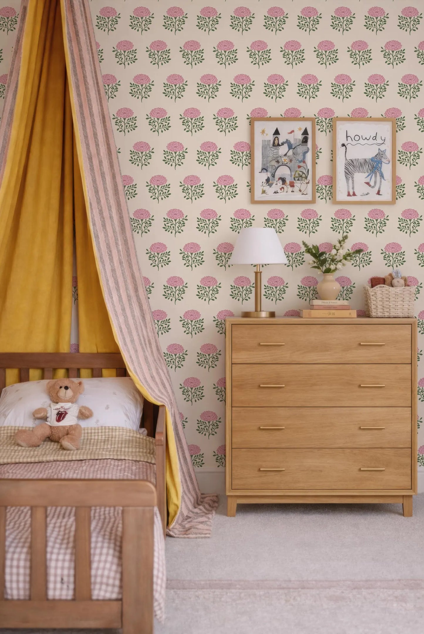a cozy and charming bedroom scene, featuring a wooden bed with a yellow and pink striped canopy, a wooden dresser, and a teddy bear. The walls are adorned with a floral wallpaper, and there are two framed pictures hanging above the dresser.