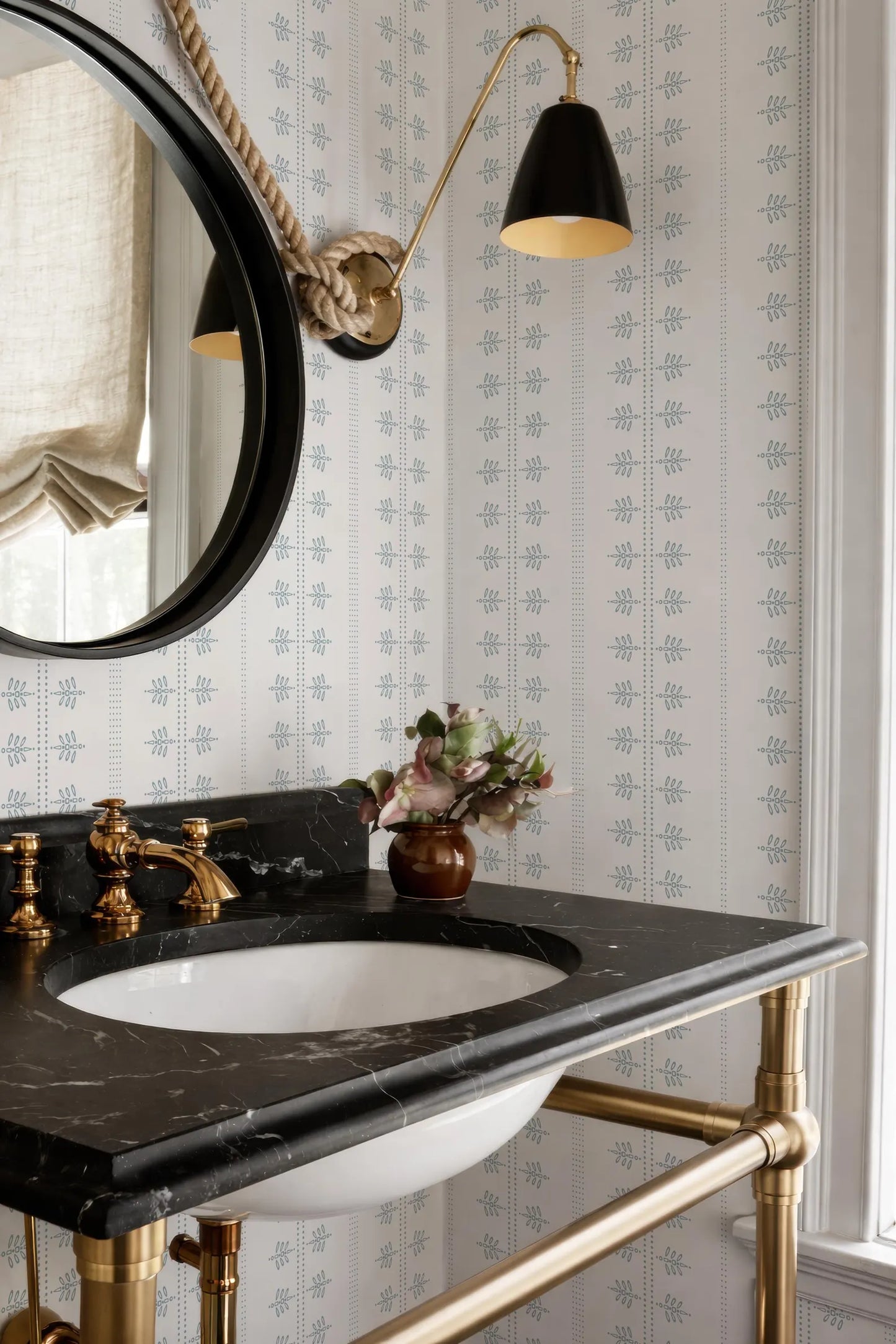 a bathroom with a black marble sink, a round mirror, and a black and gold lamp. The bathroom has a black and white patterned wallpaper, and there is a vase of flowers on the counter.