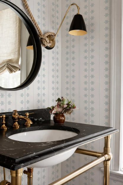 a bathroom with a black marble sink, a round mirror, and a black and gold lamp. The bathroom has a black and white patterned wallpaper, and there is a vase of flowers on the counter.
