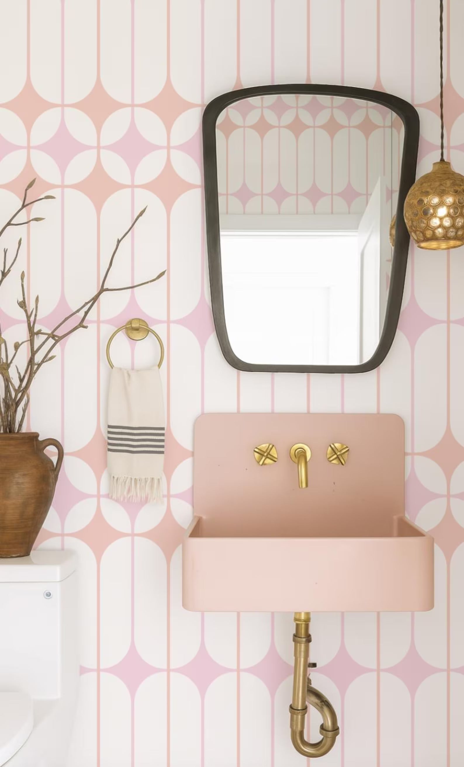a bathroom with a pink sink, a mirror, and a hanging light fixture. The walls are decorated with a pink and white geometric pattern, and there is a vase of branches on the counter.