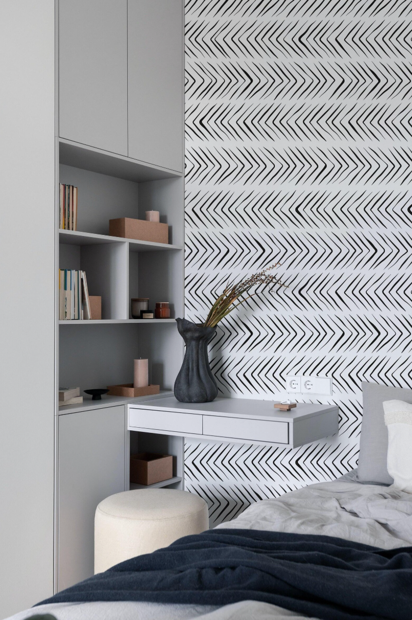 a modern bedroom with a white bookshelf, a black vase with greenery, and a gray and white zigzag patterned wallpaper.