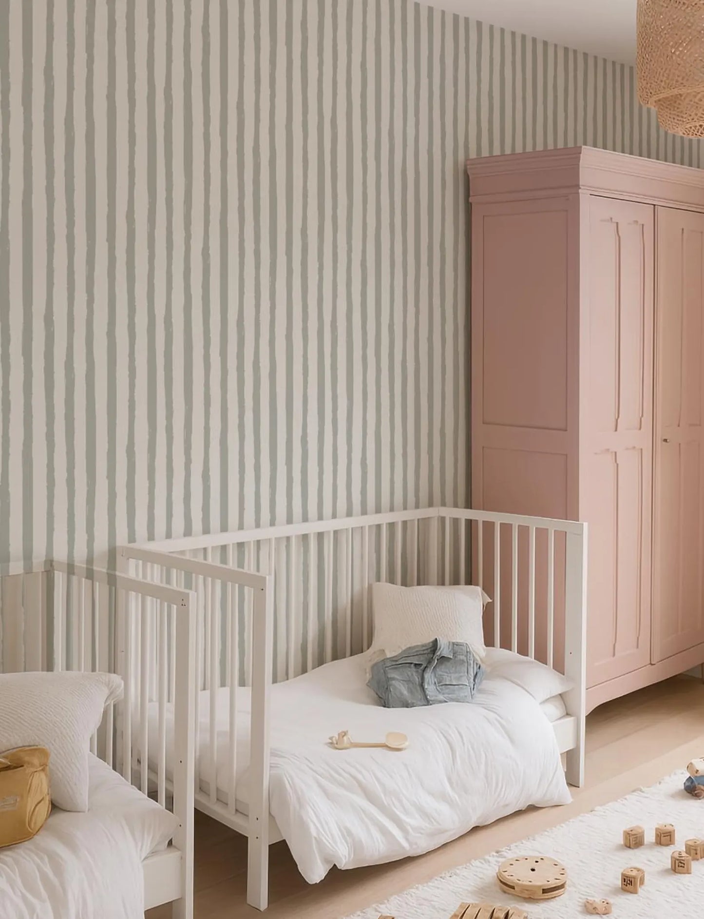 a cozy and well-organized nursery room with a white crib, a pink armoire, and a striped wallpaper.